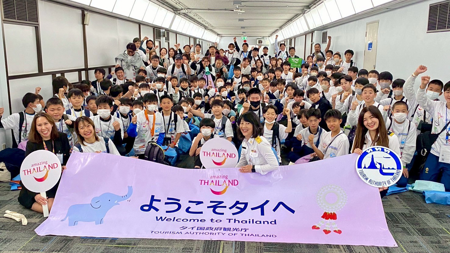 A large group of youth football players from Japan hold a “Welcome to Thailand” banner as TAT staff greet them on arrival in Bangkok.