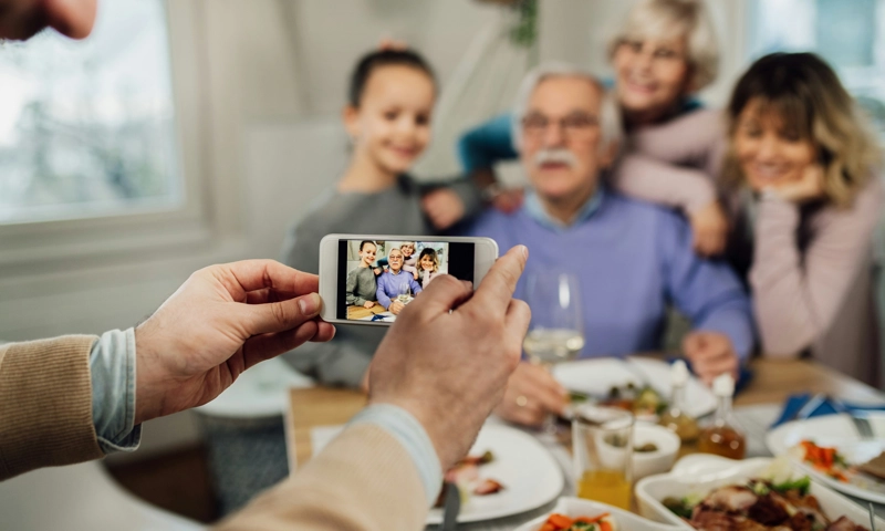 Close-up of a man using a smartphone while photographing his extended family in the dining room.