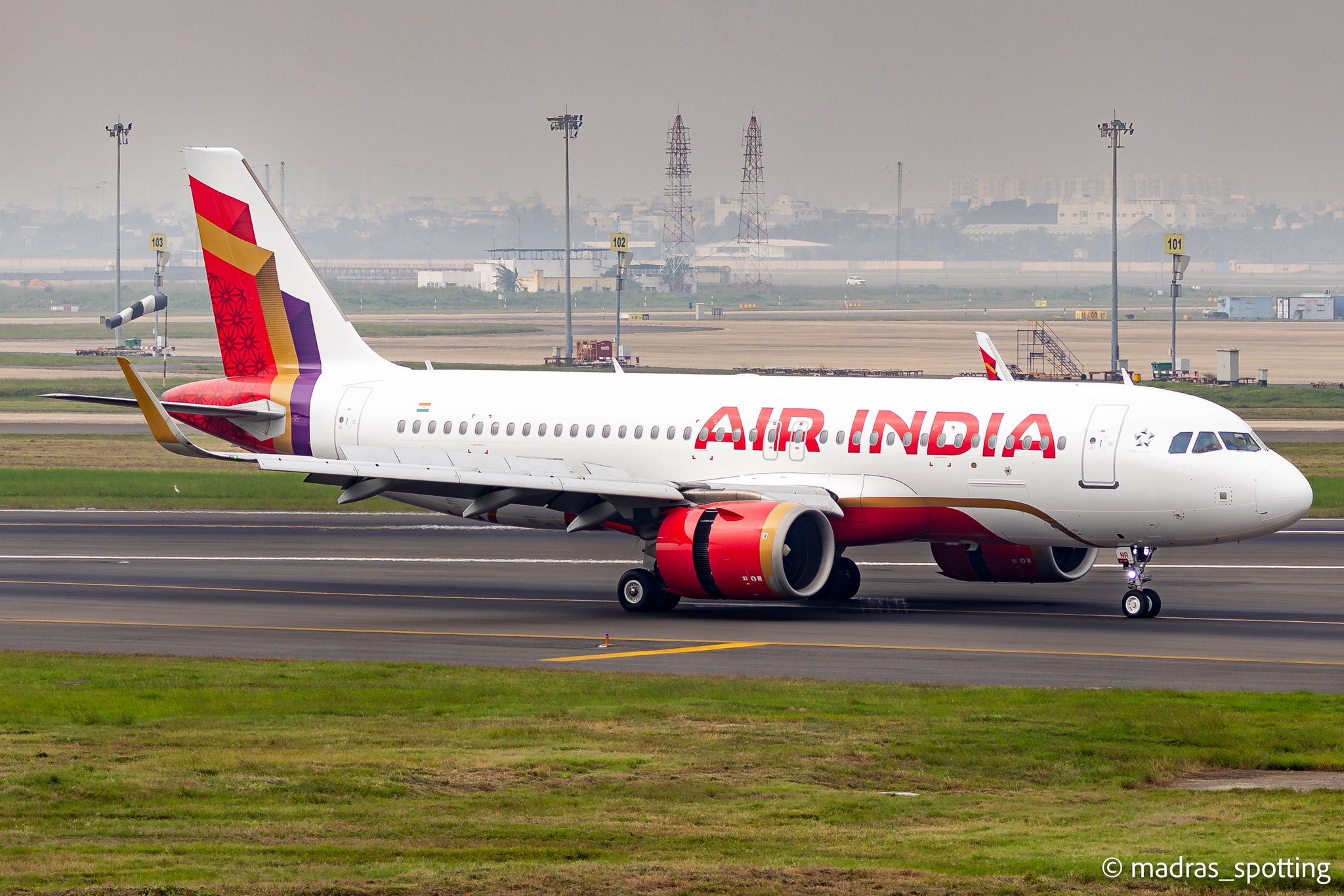 The image shows an Air India airplane on a runway at an airport. The aircraft is white with red and gold accents, and the Air India logo is visible on the fuselage. The tail features a colorful design with red, purple, and gold patterns. The background includes airport infrastructure and a hazy cityscape.