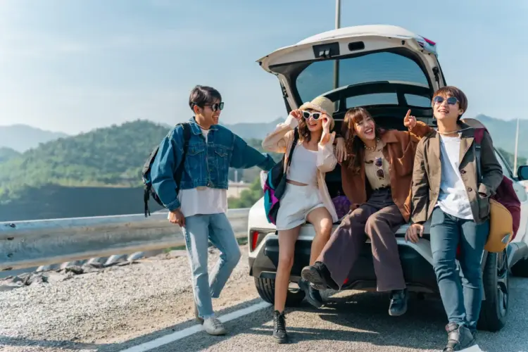 Group of young friends relaxing on the open trunk of an electric car during a summer road trip, enjoying mountain countryside views and an outdoor lifestyle.