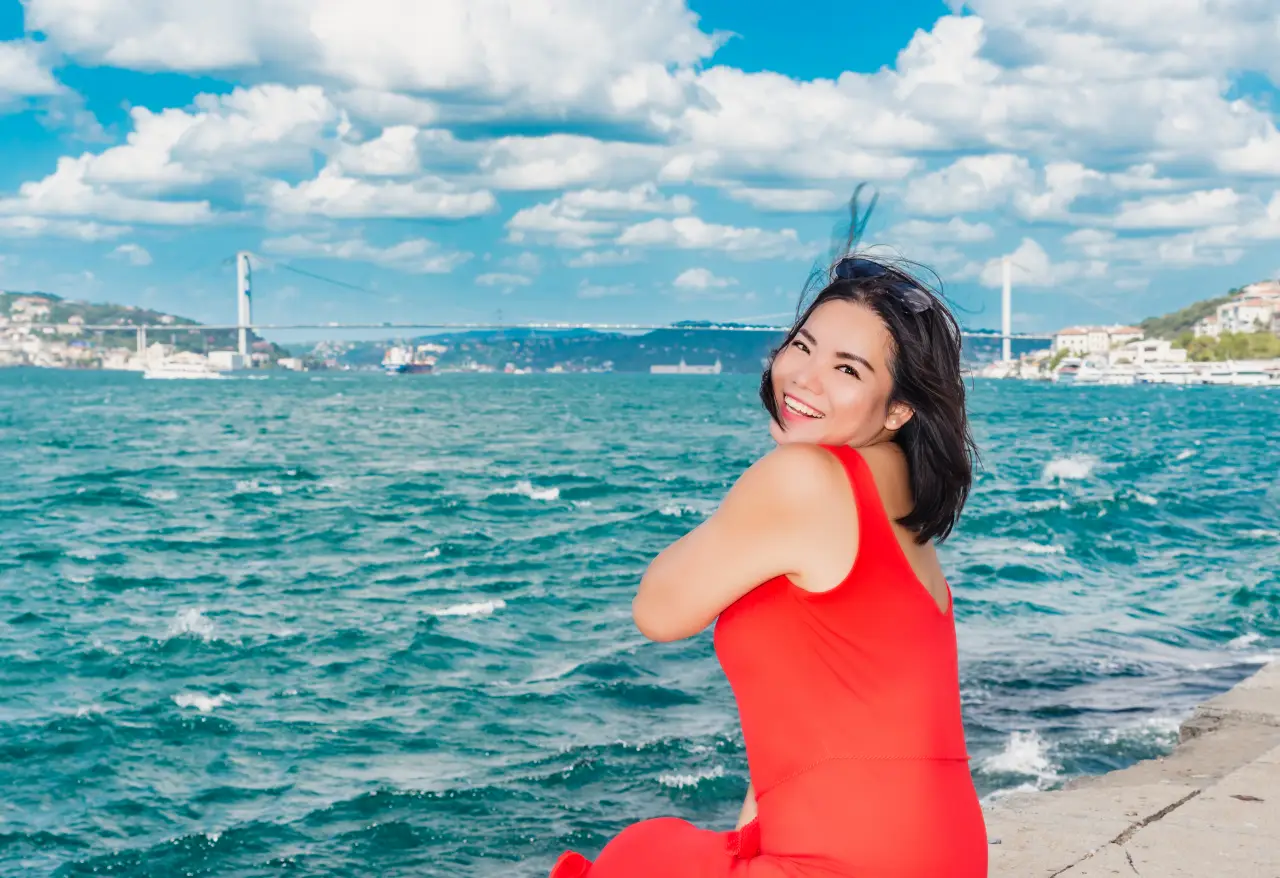 Smiling Chinese woman in a red dress sitting by the Bosphorus in Istanbul, with the bridge and sea behind her