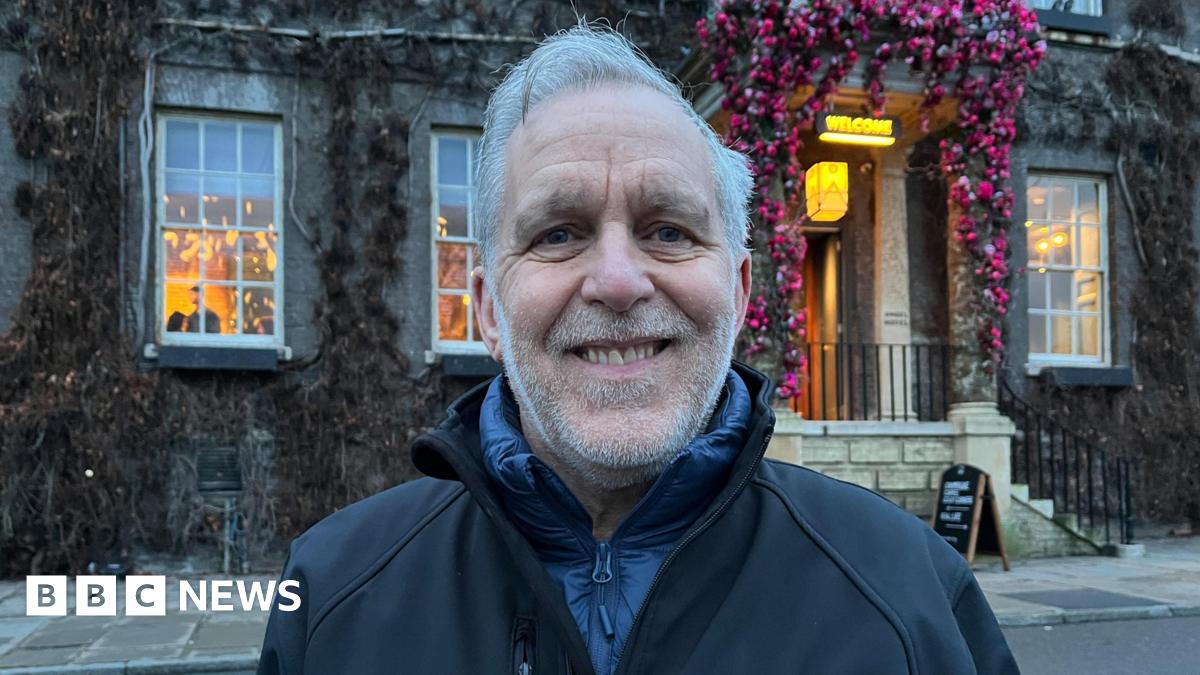 Mark Cordell wearing a black jacket labelled Our Bury St Edmunds stands outside the Angel Hotel in Bury St Edmunds. It has ivy growing over it and pink flowers around the door.