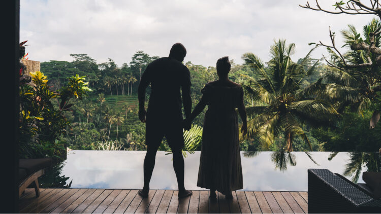 Silhouettes of two people holding hands on a deck overlooking a tropical infinity pool and lush green jungle.