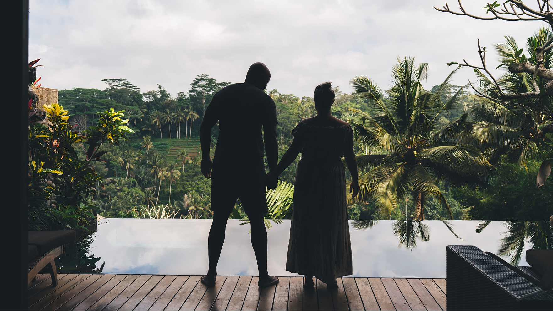 Silhouettes of two people holding hands on a deck overlooking a tropical infinity pool and lush green jungle.