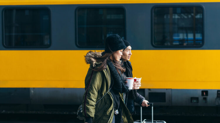 Couple in winter coats walking with luggage along a platform beside a yellow train.