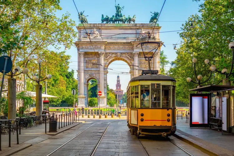 Yellow tram passing through the Arch of Peace in Milan on a sunny day with green trees.