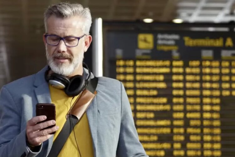 Business traveler looks at his phone in an airport. There is a flight board in the background.