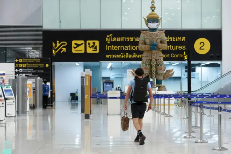 Traveler walking through a quiet international departures hall at a Thailand airport with a masked guardian statue and signage overhead