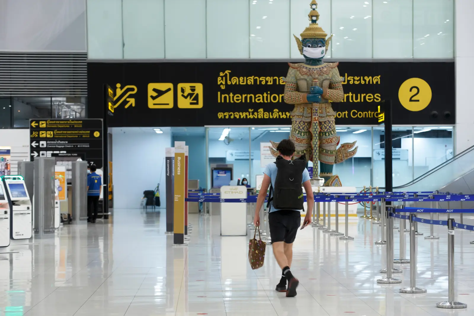 Traveler walking through a quiet international departures hall at a Thailand airport with a masked guardian statue and signage overhead