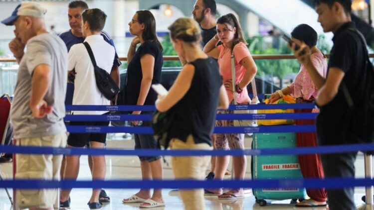 Several people in colourful summer clothes queue around blue barriers at a bright airport forecourt.