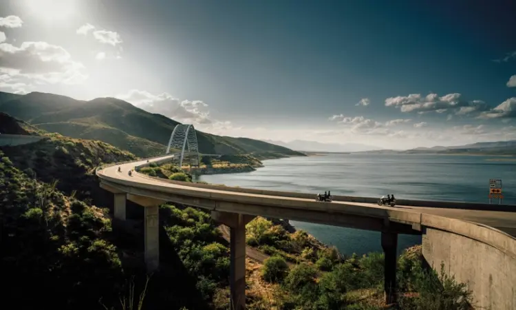 A scenic motorcycle road trip crossing a bridge over a large body of water, surrounded by mountainous terrain.