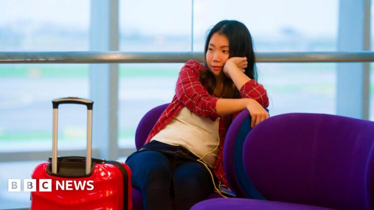 Young woman in red checked shirt rests her chin on her hand. She is sitting in an airport waiting room with a red case next to her