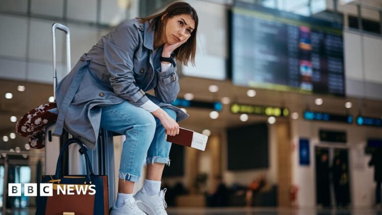 Young woman wearing blue jeans, a grey trenchcoat and white trainers sits on her suitcase at an airport, clutching her passport and ticket