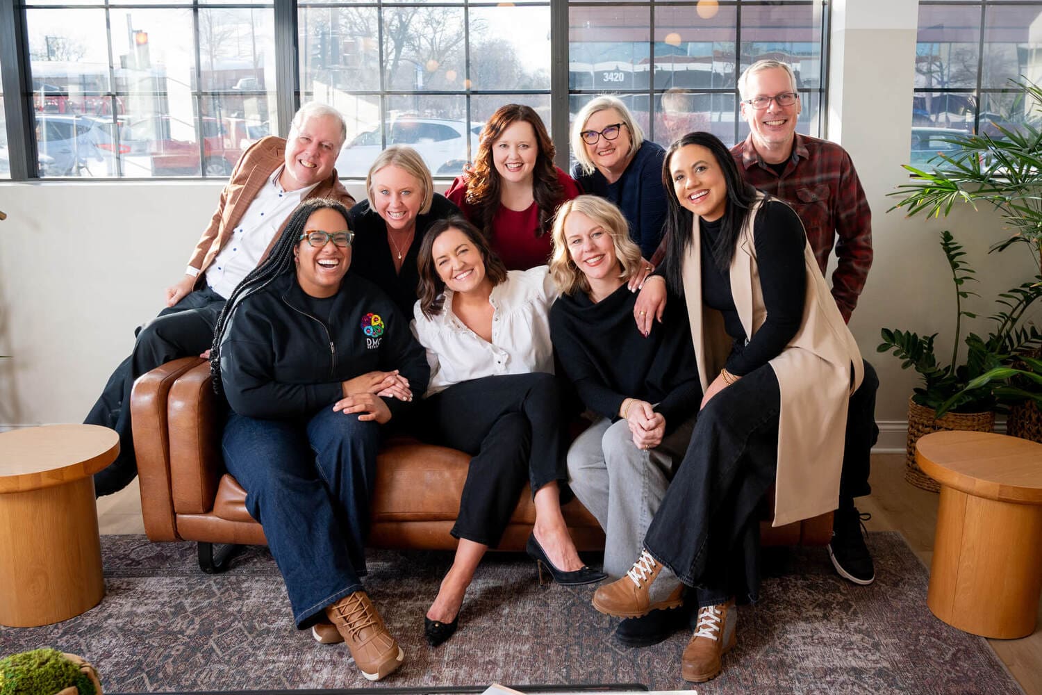 A group of nine smiling colleagues, men and women of various ages, pose together in a bright, modern office space. Some are seated on a brown leather sofa while others lean in from behind. They are dressed in smart-casual attire, and the room features large windows and indoor plants, creating a warm and professional atmosphere.