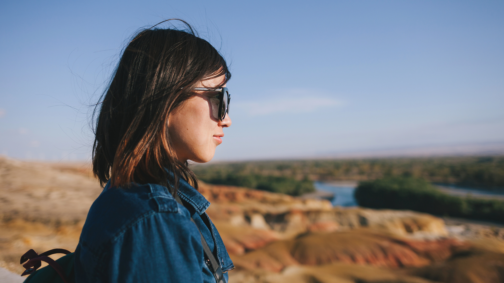 Side-profile image of a woman wearing sunglasses and a blue shirt, standing outdoors and looking over a sunlit canyon landscape with layered earth tones, greenery, and a winding river in the distance.