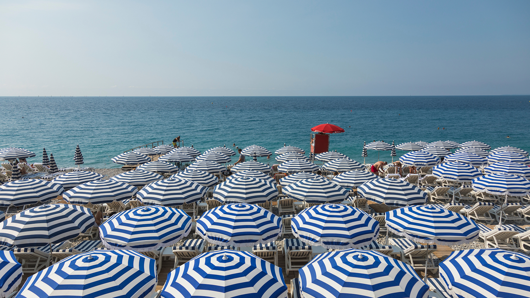 Wide view of a beach filled with rows of blue-and-white striped umbrellas and lounge chairs facing a calm blue sea, with a red lifeguard stand near the shoreline under a clear sky.