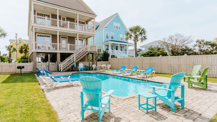 Exterior view of a large coastal vacation rental with two covered balconies overlooking a rectangular private swimming pool. The pool is surrounded by a paved patio, blue lounge chairs, turquoise Adirondack chairs, a white privacy fence, palm trees, and neighboring beach houses in the background.