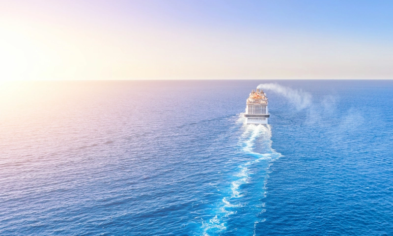 large cruise ship sailing through deep blue ocean waters at sunset or sunrise, leaving a distinct white wake behind it.